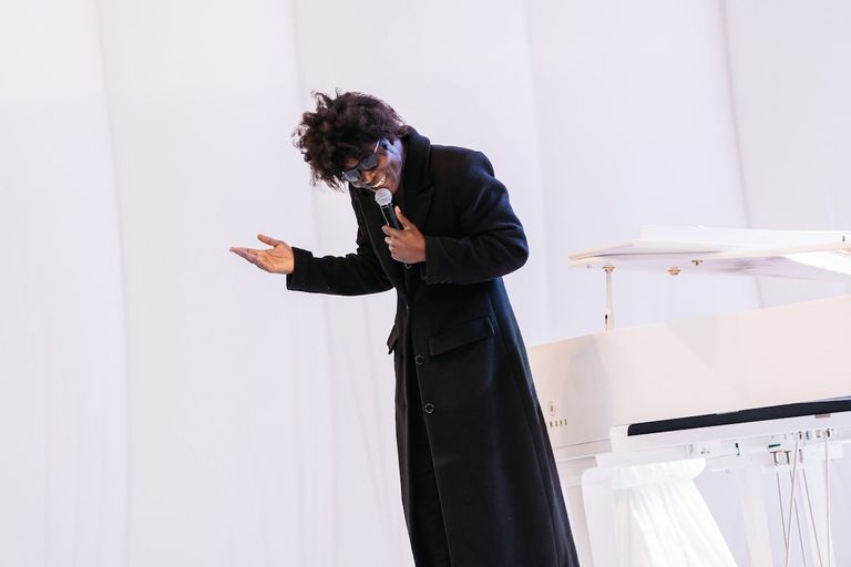 Musician Benjamin Clementine performs in a long black coat and sunglasses, holding a microphone next to a white grand piano.